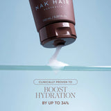 A close-up of a brown squeeze tube of Nak Hair leave-in conditioner with white cream being dispensed, set against a light blue background with text below.