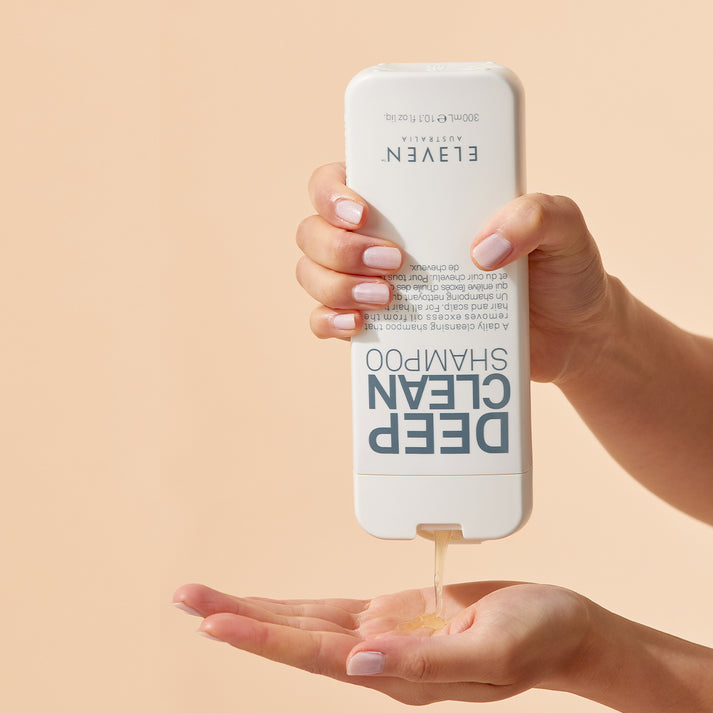 Hand holding a bottle of Eleven Deep Clean Shampoo with shampoo being poured into another hand on a beige background