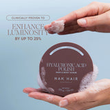 A person's hands holding a round brown container of NAK HAIR Hyaluronic Acid Polish Hair & Body Scrub, with white foam around the container and one hand lifting a piece of scrub. Text on the left reads 'CLINICALLY PROVEN TO ENHANCE LUMINOSITY BY UP TO 25%'.